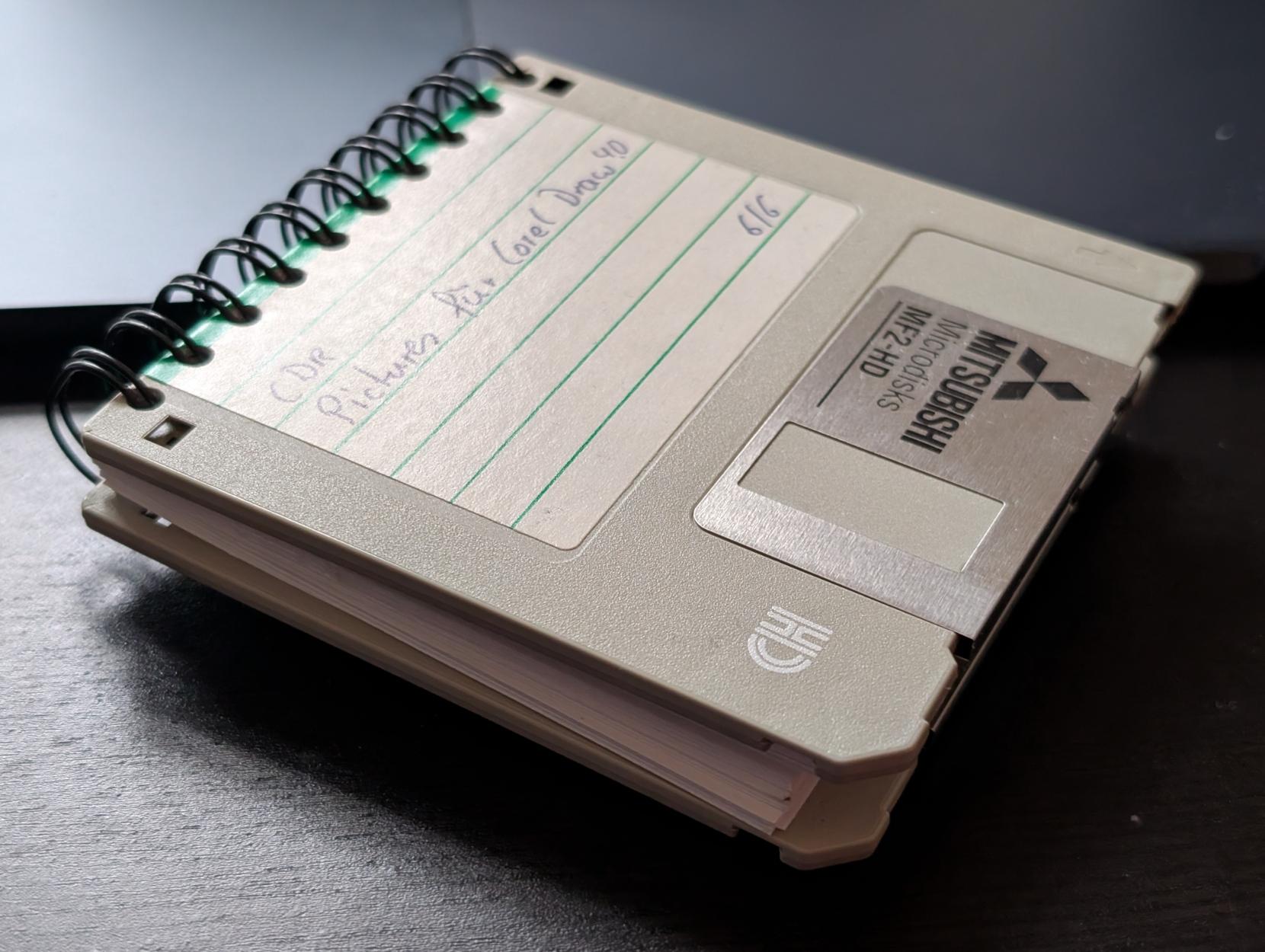two grey 3 1/2 inch floppy disks bound as lid and base into a square paper notebook. the hand written labels indicate they had been used for pictures for corel draw 4.0